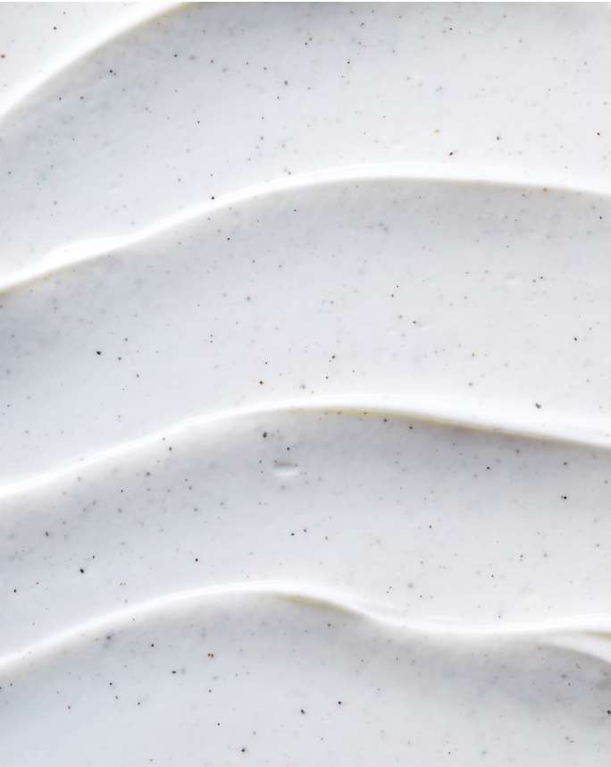 Close-up of a thick, white cosmetic cream with tiny dark granules, swirled to create a textured, wave-like surface pattern.