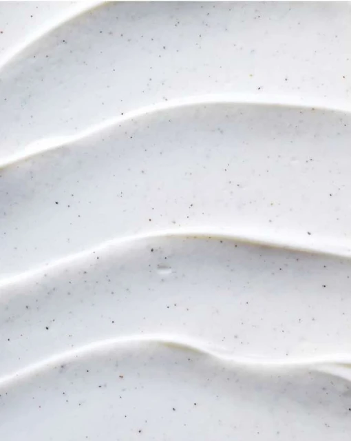 Close-up of a thick, white cosmetic cream with tiny dark granules, swirled to create a textured, wave-like surface pattern.