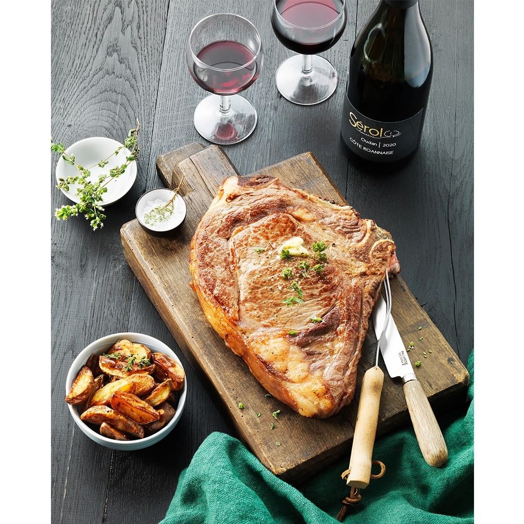 Overhead shot of ribeye steak on a wooden board, garnished with herbs, accompanied by wine, potatoes, knife, and green cloth on a dark table.
