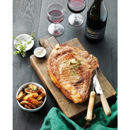 Overhead shot of ribeye steak on a wooden board, garnished with herbs, accompanied by wine, potatoes, knife, and green cloth on a dark table.