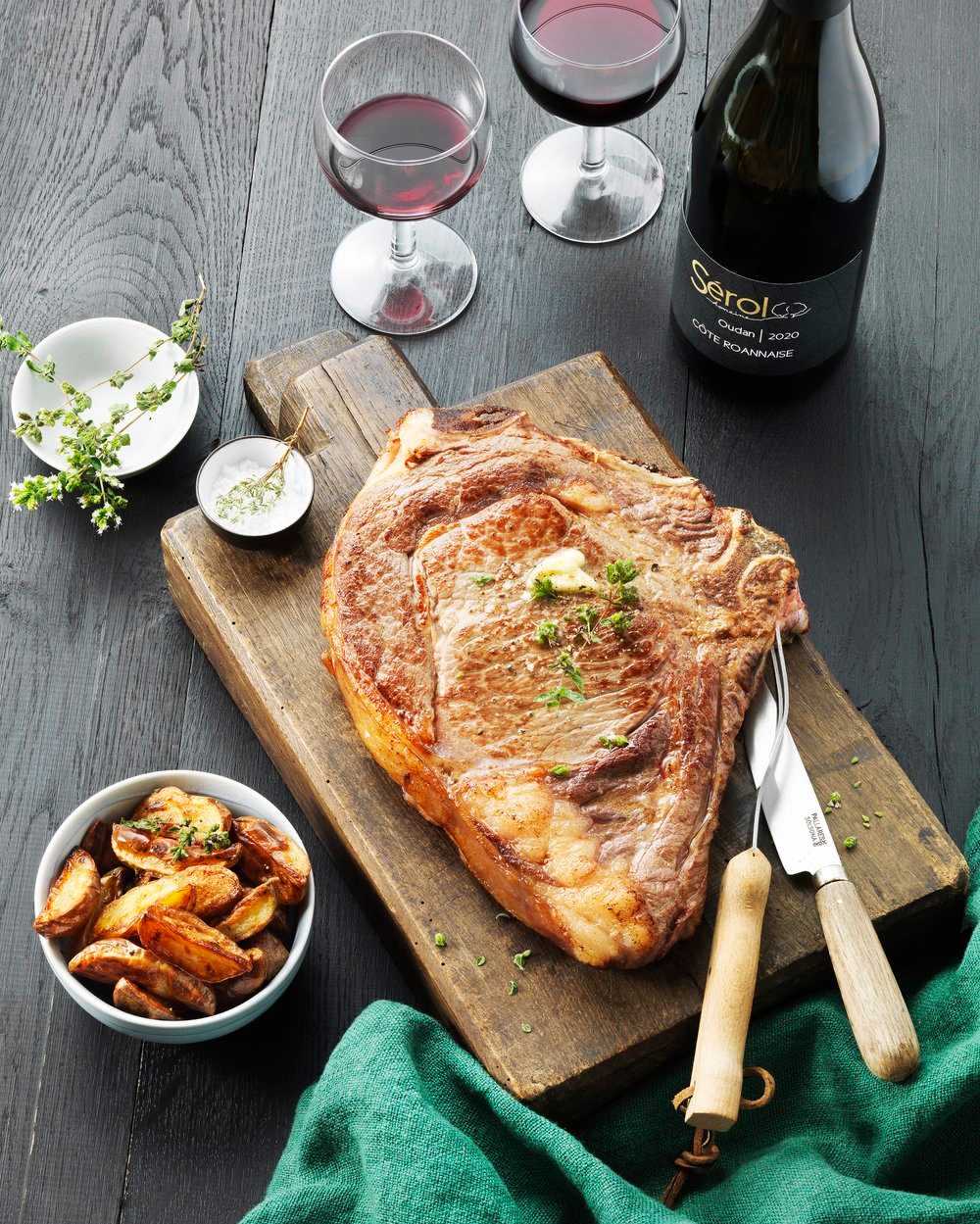 Overhead shot of a cooked steak on a wood board, served with red wine, potatoes, spices, and garnished with greens on dark wood table.