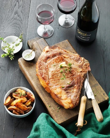 Overhead shot of a cooked steak on a wood board, served with red wine, potatoes, spices, and garnished with greens on dark wood table.