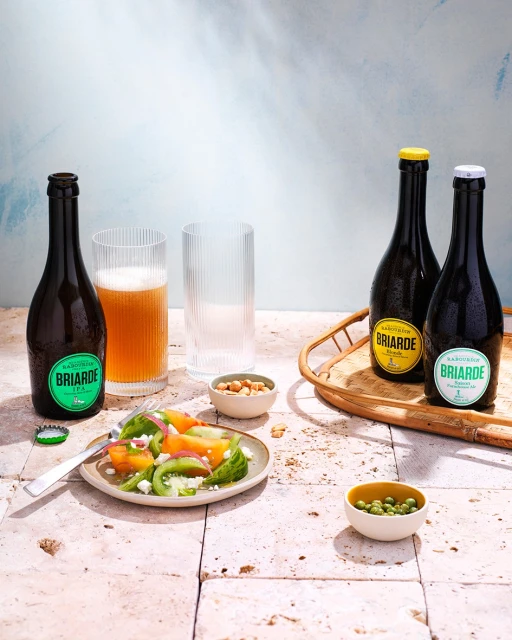 Still life featuring Briarde beer bottles, salad, and snacks on a stone table against a textured backdrop, illuminated by studio lighting.