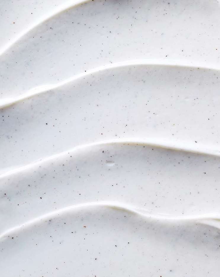 Close-up of white cream with dark specks, showing a wavy texture. The cream appears thick and smooth, creating a rippled pattern.