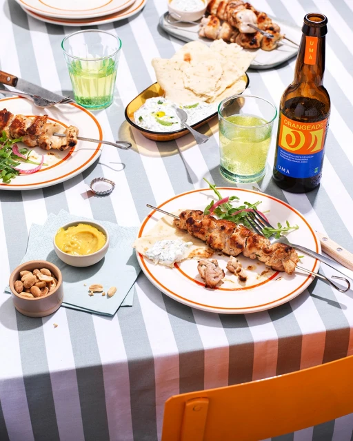 Overhead shot of plates with chicken skewers, pita bread, dips, and drinks on a striped tablecloth, creating a vibrant food display.