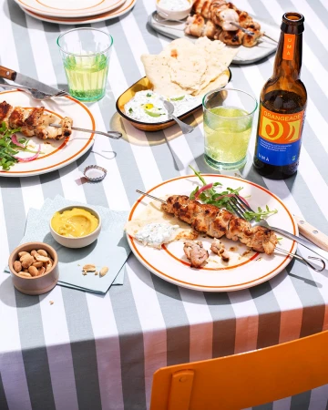 Overhead shot of plates with chicken skewers, pita bread, dips, and drinks on a striped tablecloth, creating a vibrant food display.