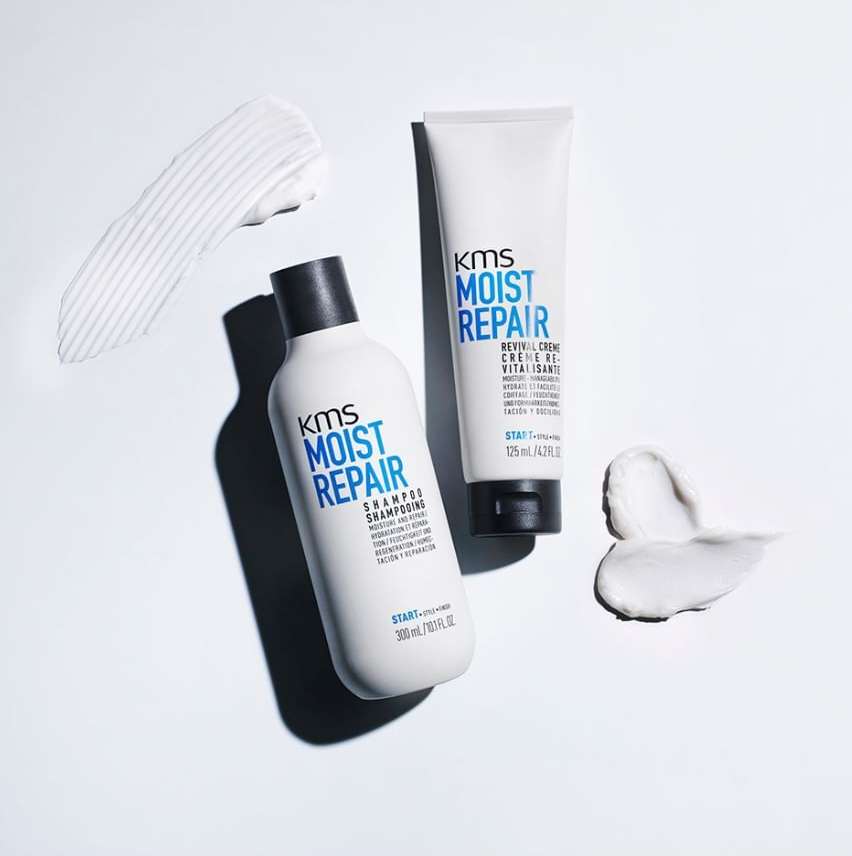 Two white bottles of KMS Moist Repair shampoo and revival creme are staged with white cream smears against a white background.
