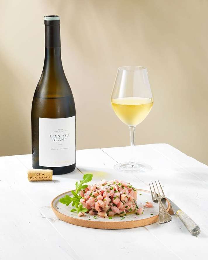 Still life shot of Anjou Blanc wine bottle, wine glass, cork, fork, knife, and plate of food with garnish on a white table.