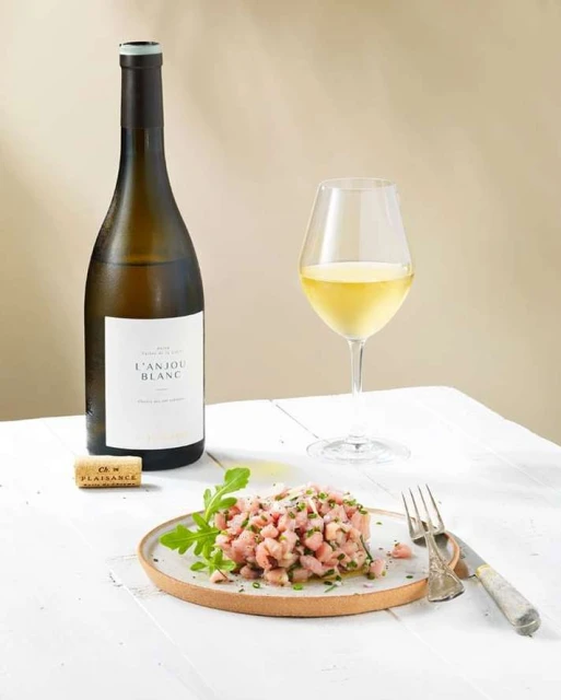 Still life shot of Anjou Blanc wine bottle, wine glass, cork, fork, knife, and plate of food with garnish on a white table.