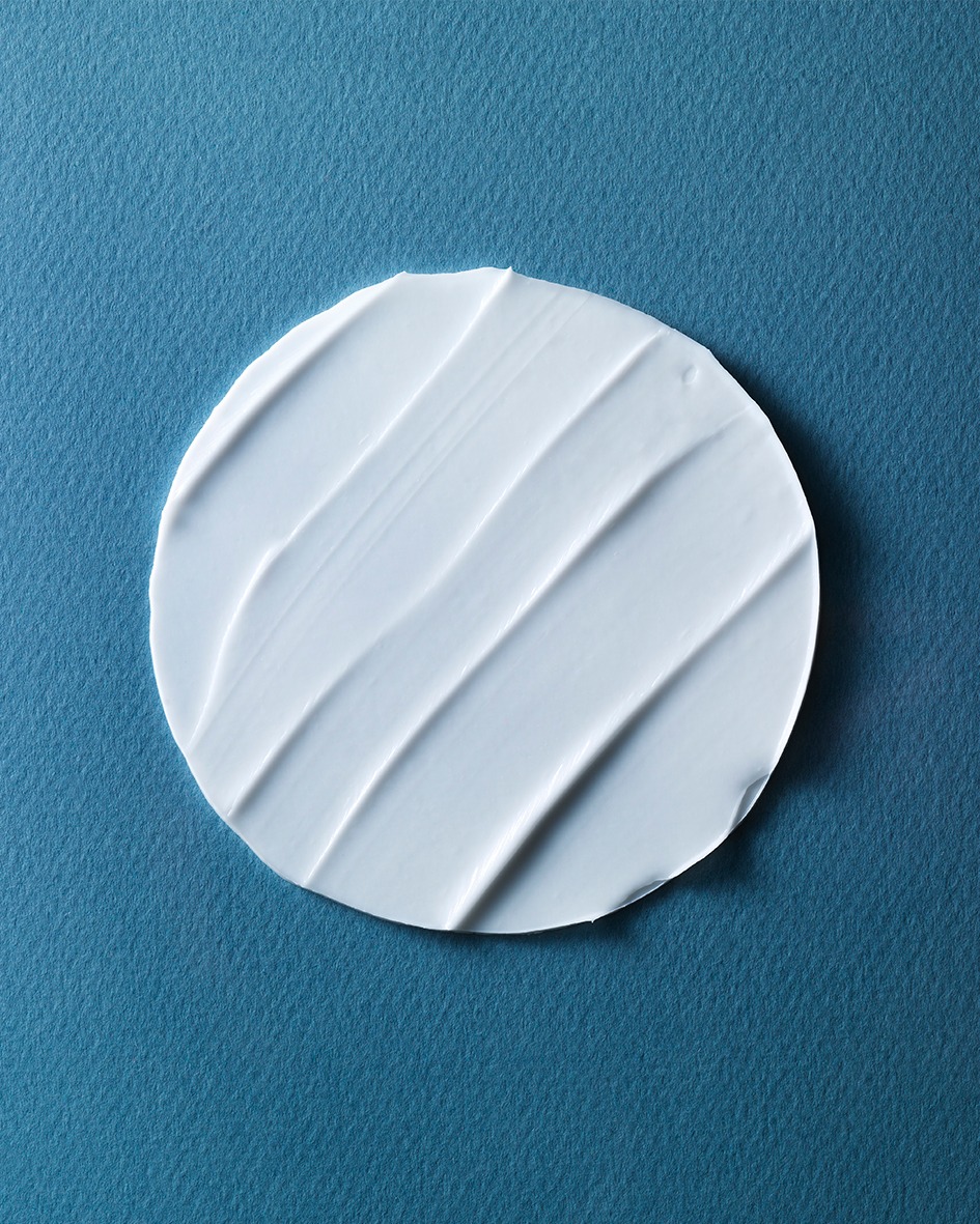 Close-up of a circular swatch of white cream or lotion with textured lines, set against a solid light blue background.