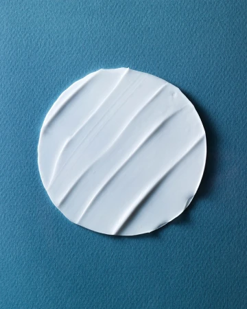 Close-up of a circular swatch of white cream or lotion with textured lines, set against a solid light blue background.