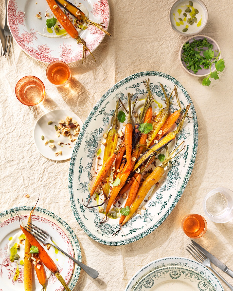 Overhead shot of roasted carrots with nuts on patterned plates, surrounded by drinking glasses and cutlery on a beige tablecloth.