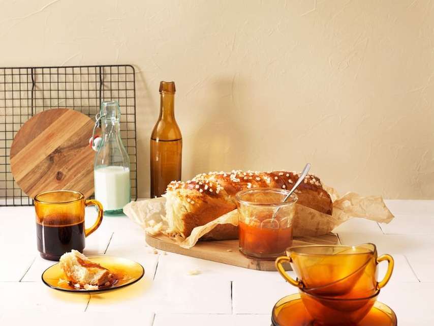 Still life photograph of a brioche loaf, jam, amber glassware, and a bottle of milk, arranged on a white wooden table against a beige backdrop.