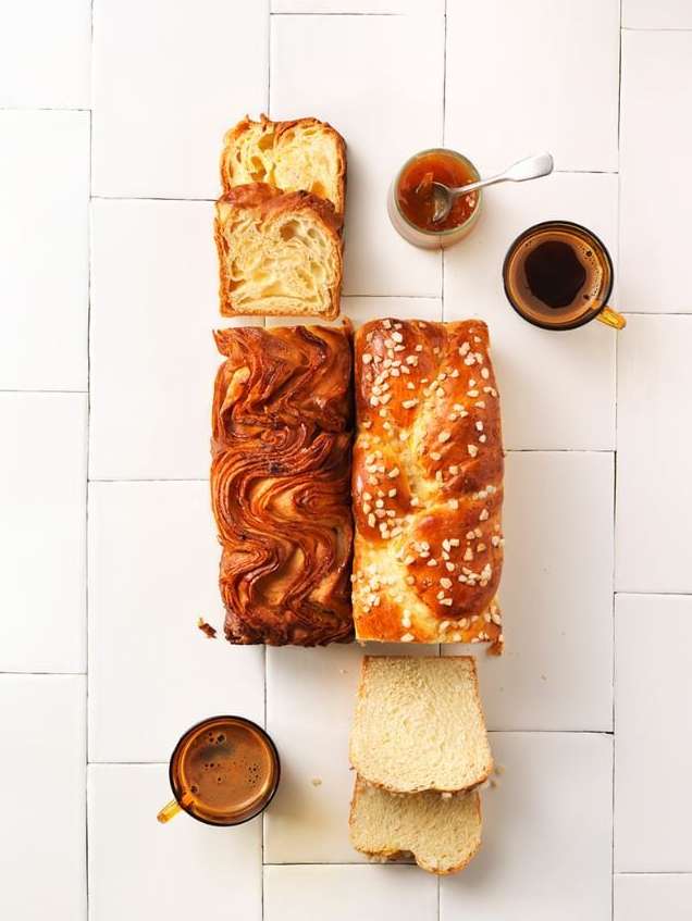 Overhead shot of arranged baked bread loaves, bread slices, coffee cups, and jam on a white tile surface, showcasing culinary composition.