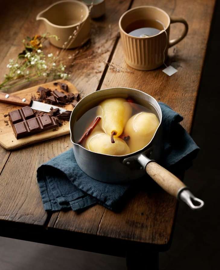 Pears in syrup sit in a metal pan on a rustic wooden table next to a mug of tea and dark chocolate.