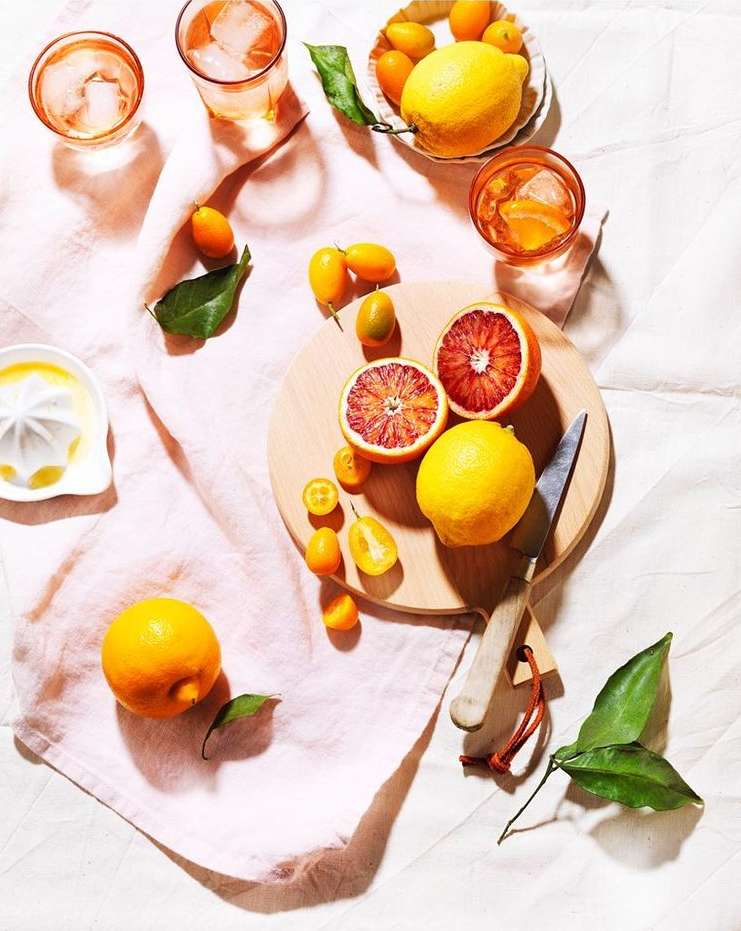 Overhead shot of blood oranges, lemons, and kumquats on a wooden cutting board and in glasses, arranged on a textured pink linen.