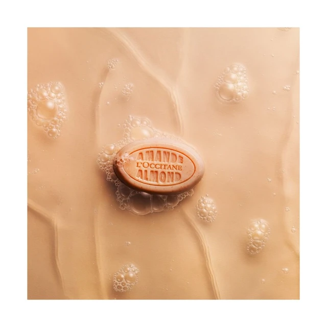 Oval almond soap bar with logo on tan, rippled liquid surface with tiny bubbles; beauty or skincare product shot.