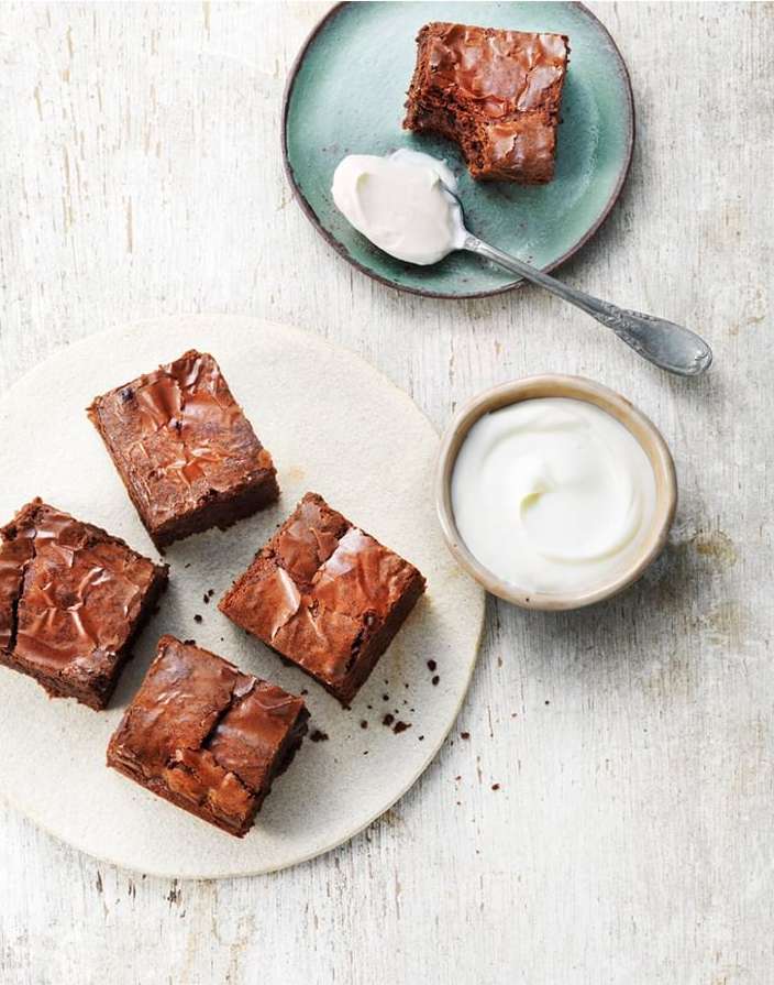 Overhead shot of brownie squares arranged on plates with a spoonful and bowl of white cream on a light, weathered wood surface.