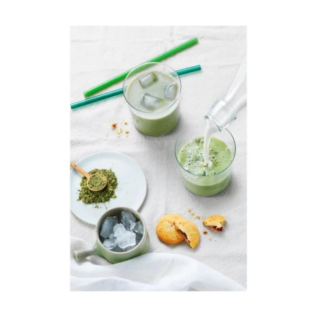 Iced matcha latte drinks with cookies. Milk is poured into a matcha-filled glass with ice, next to matcha powder and cookies.