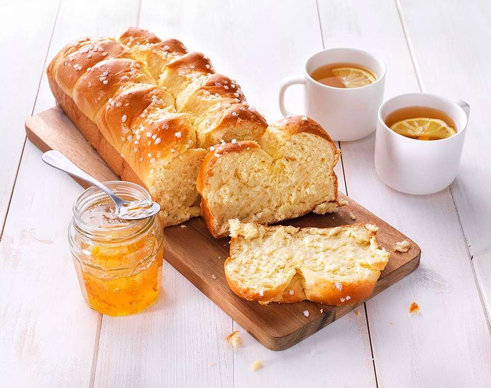 Overhead shot of a sliced braided loaf of bread with pearl sugar next to a jar of marmalade and two cups of tea on a wooden board.
