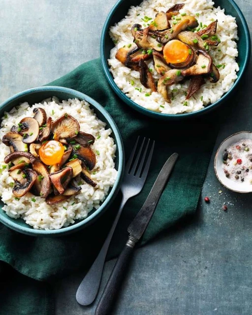 Two bowls of rice topped with sauteed mushrooms, green onions, and a fried egg yolk on a teal background with silverware.