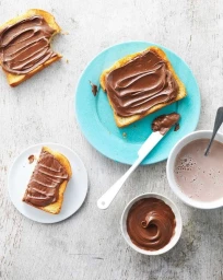 Overhead food photography of chocolate spread on toast slices, a blue plate, a small white bowl, a white mug, and a knife on a grey wooden surface.