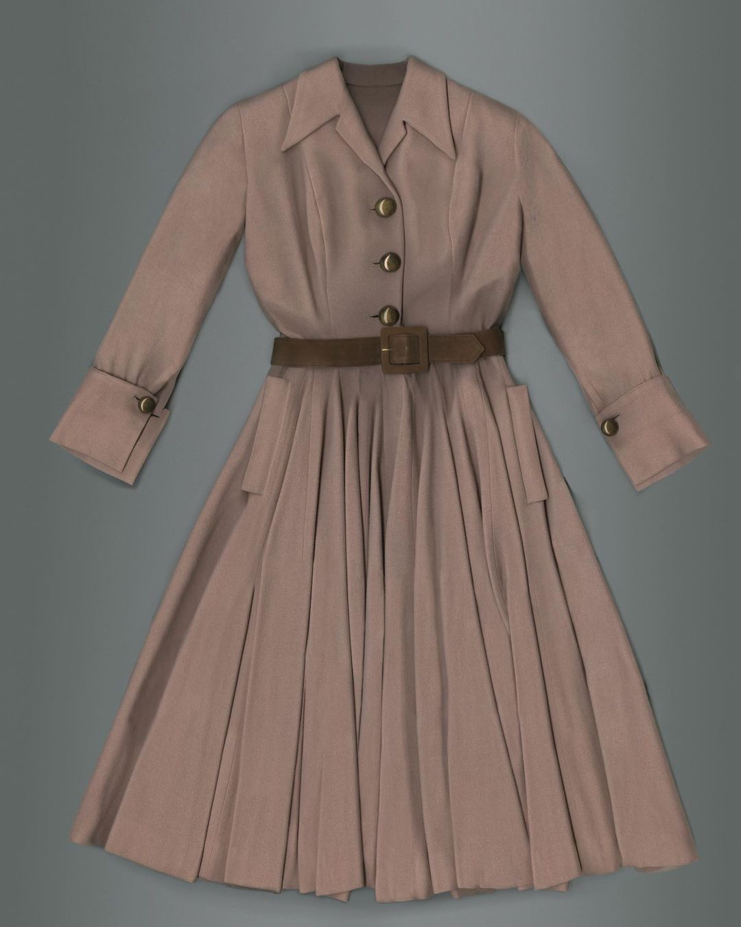 Tan dress with a collared neck, long sleeves with buttoned cuffs, a brown belt, and a pleated skirt on a gray background.
