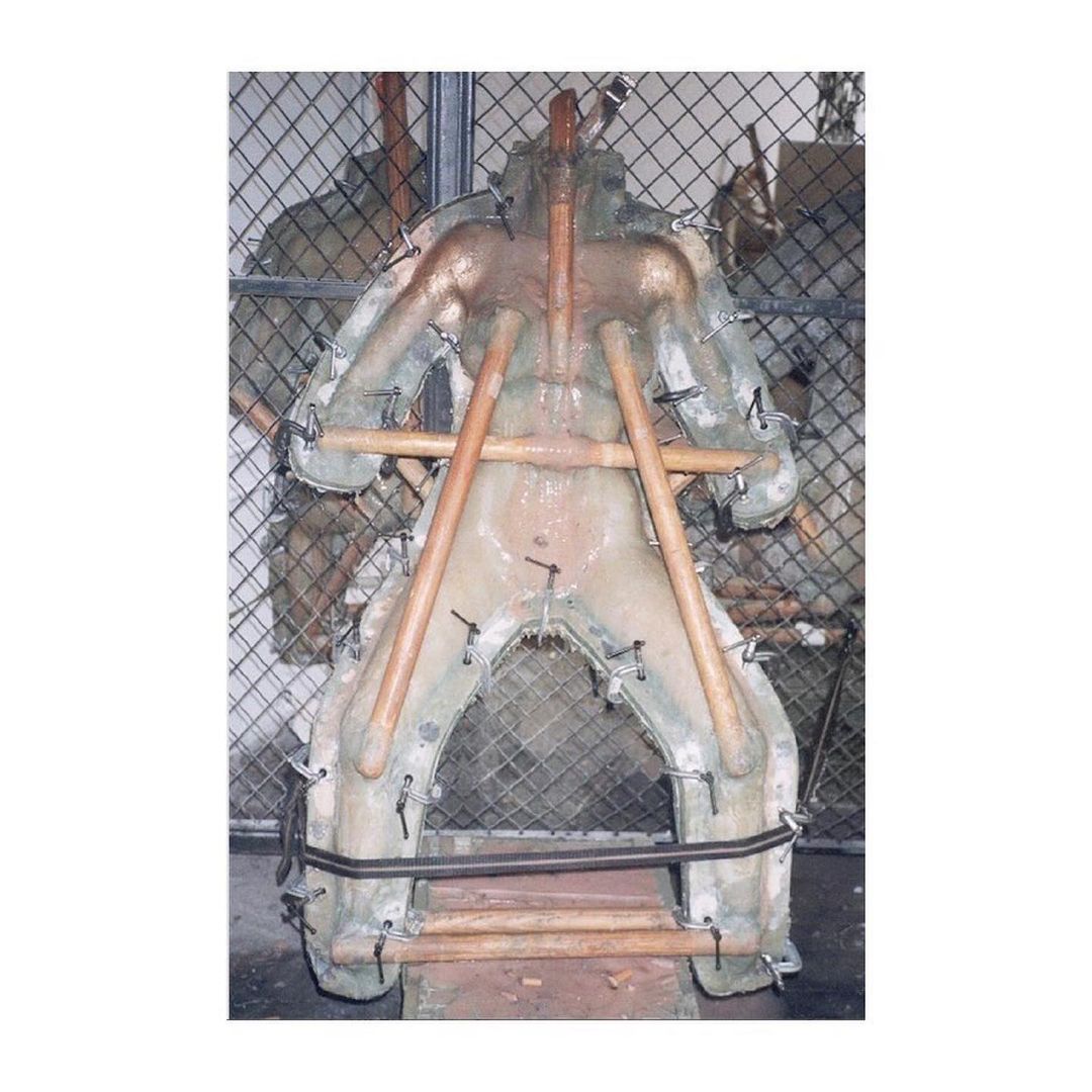 Life-size torso body mold, made of a light yellow material, is held together with wood supports and metal pins. Displayed indoors.