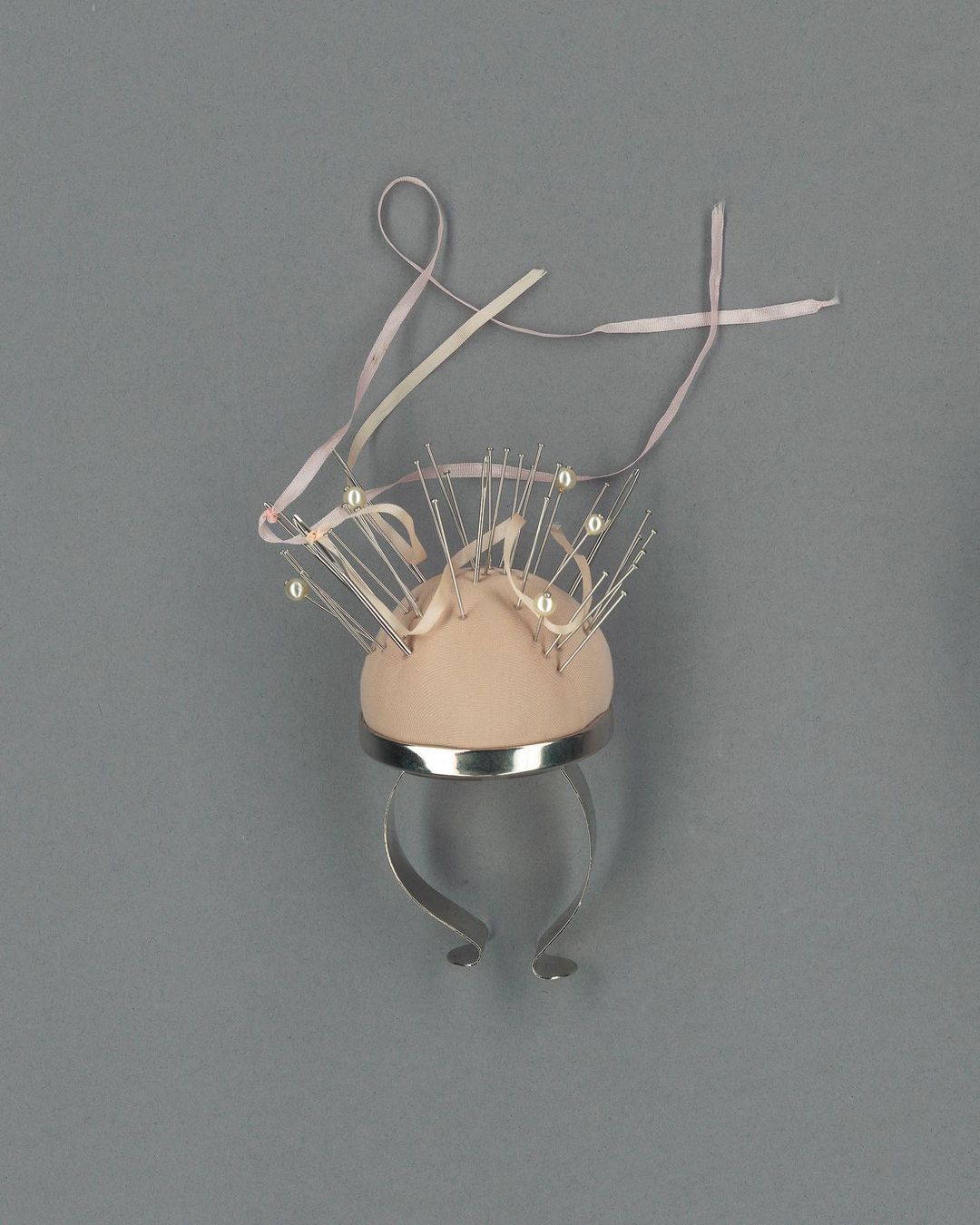 Unique sculptural design featuring a domed, soft-pink form adorned with pearl-tipped pins and a ribbon, set against a grey background.