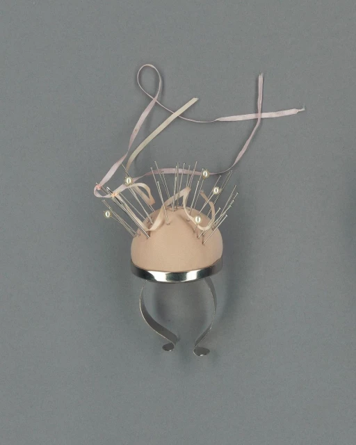 Unique sculptural design featuring a domed, soft-pink form adorned with pearl-tipped pins and a ribbon, set against a grey background.