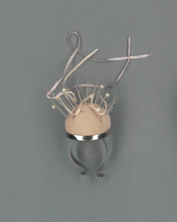 Unique sculptural design featuring a domed, soft-pink form adorned with pearl-tipped pins and a ribbon, set against a grey background.