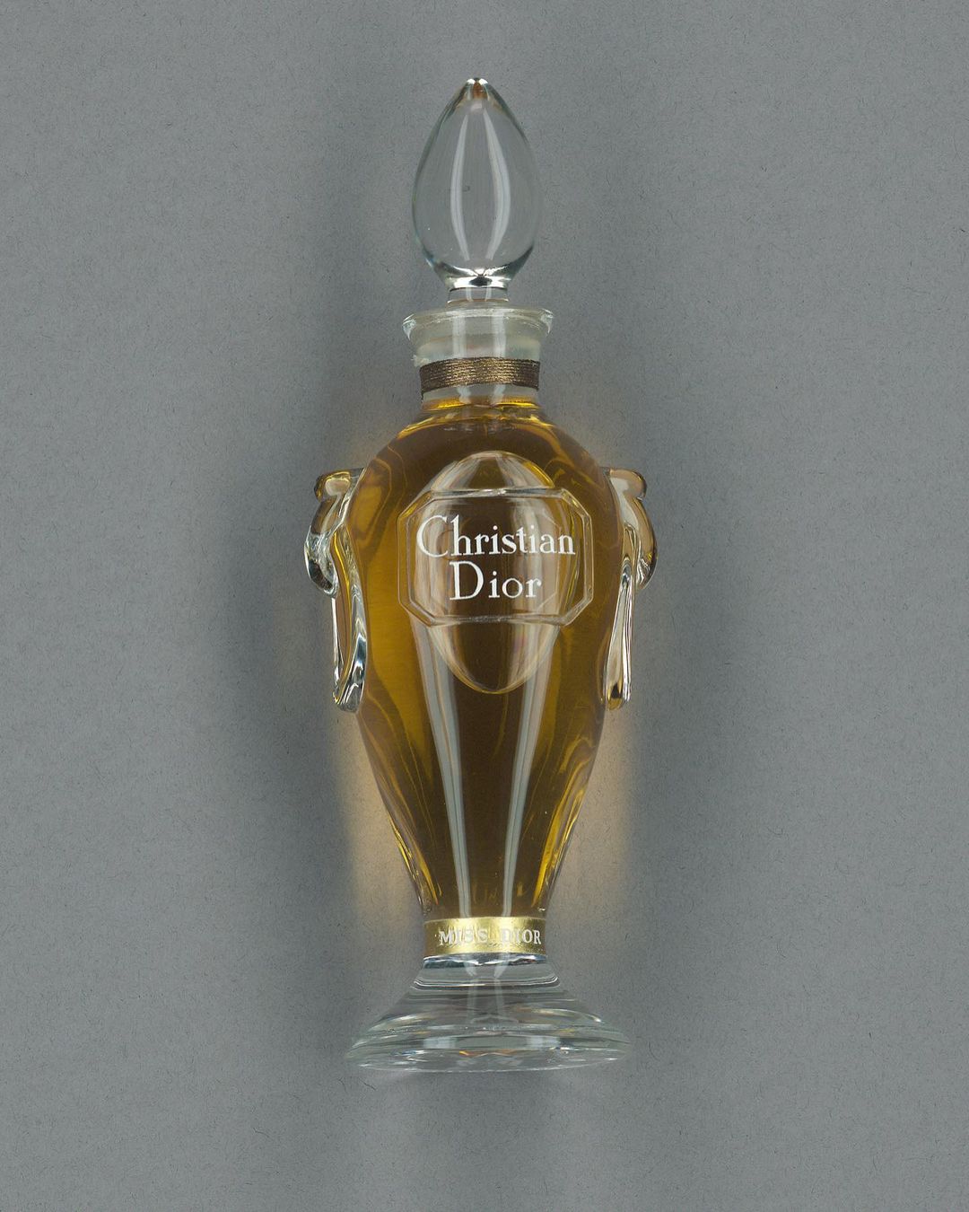 Elegant glass perfume bottle filled with golden liquid. The bottle has a clear, decorative stopper and a label with the brand name.