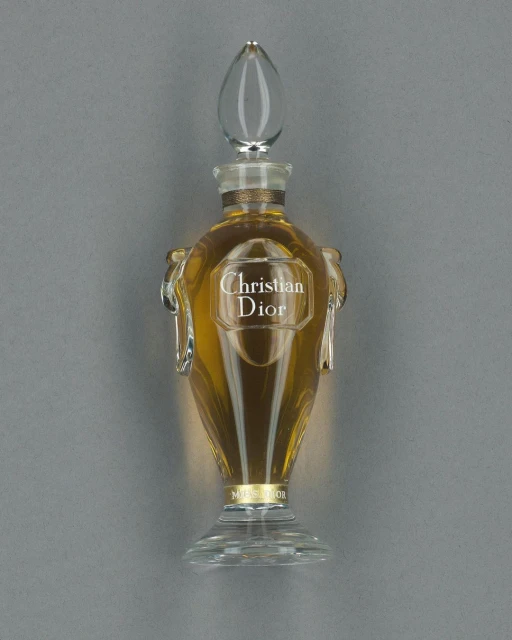 Elegant glass perfume bottle filled with golden liquid. The bottle has a clear, decorative stopper and a label with the brand name.