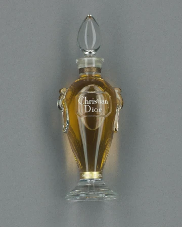 Elegant glass perfume bottle filled with golden liquid. The bottle has a clear, decorative stopper and a label with the brand name.