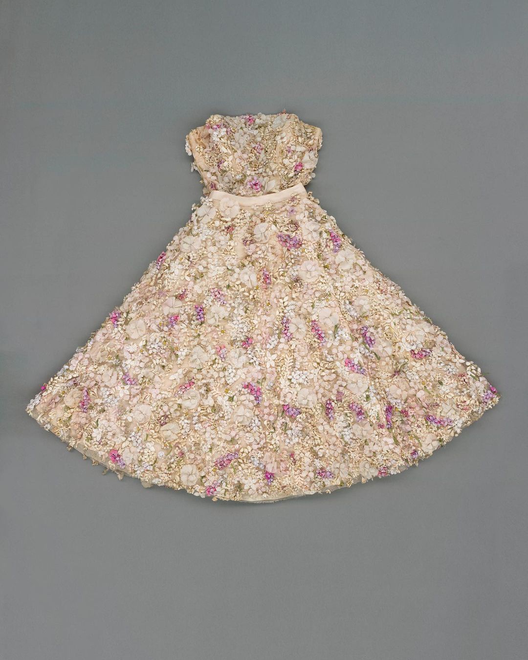 Strapless dress with floral embellishments in white, cream, and pink hues, photographed on a neutral grey background.
