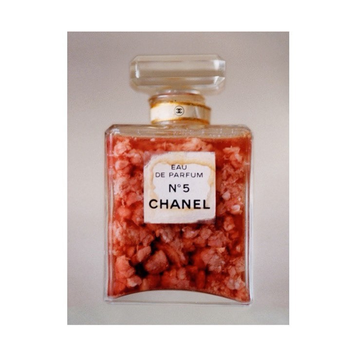 Square-shaped Chanel No. 5 perfume bottle filled with coral colored stones against a plain background in a studio shot.