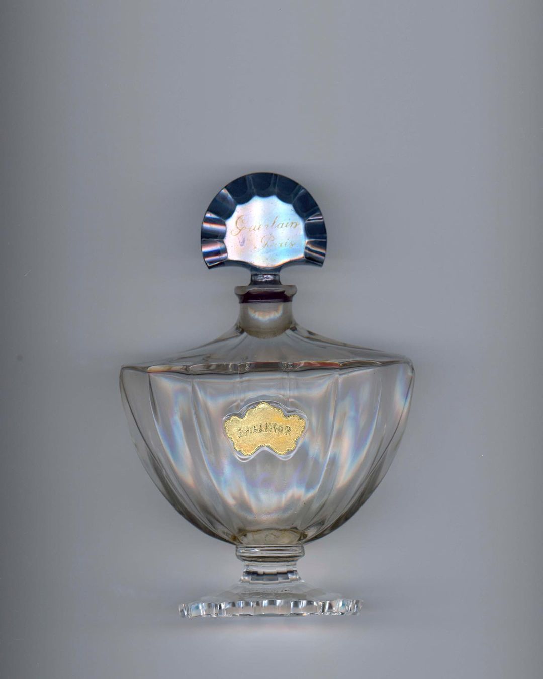 Elegant clear glass perfume bottle with a silver top and a gold label on a neutral background, showcasing vintage design.