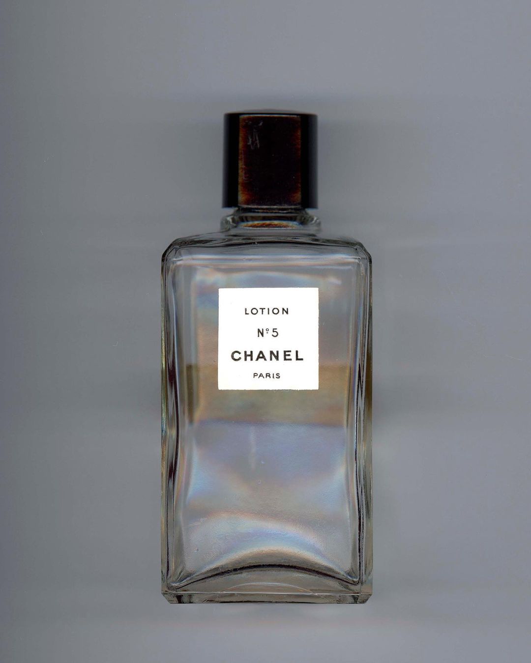 Chanel No. 5 lotion bottle on a gray surface. Clear rectangular glass container with brown top and white label. Minimalist beauty shot.