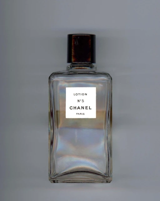 Chanel No. 5 lotion bottle on a gray surface. Clear rectangular glass container with brown top and white label. Minimalist beauty shot.