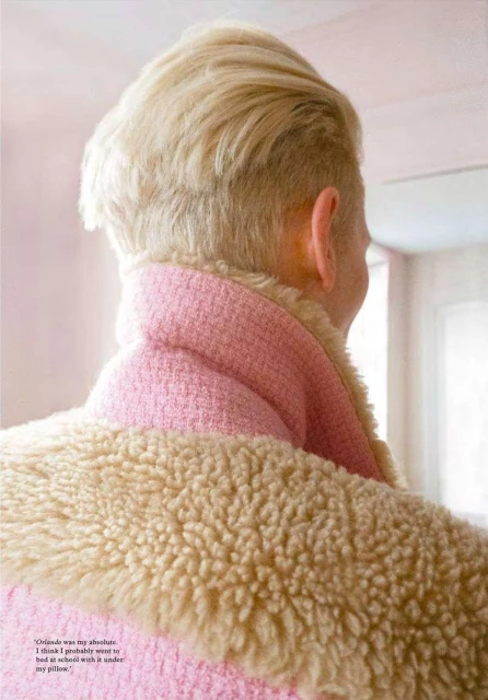 An adult female with a blond undercut is shown from the back, wearing a pink jacket with a cream-colored fur collar, in a bright, indoor setting.