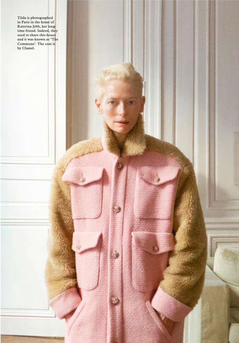Fashion editorial shot of a fair-skinned adult female with short blonde hair, wearing a pink coat with brown fur sleeves indoors.