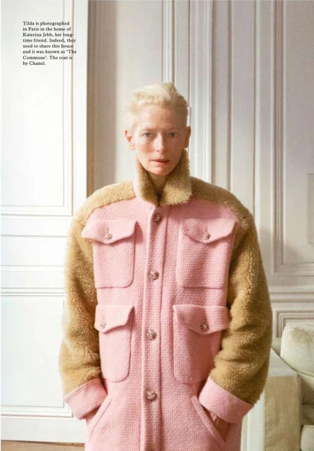 Fashion editorial shot of a fair-skinned adult female with short blonde hair, wearing a pink coat with brown fur sleeves indoors.