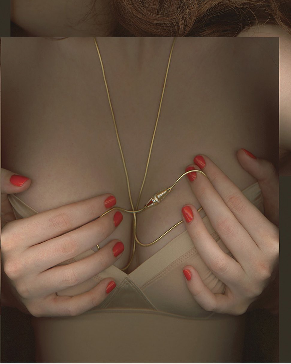 Close-up shot of a woman with red nails wearing a bra and a golden pendant with a geometric connector, hands resting on her chest.