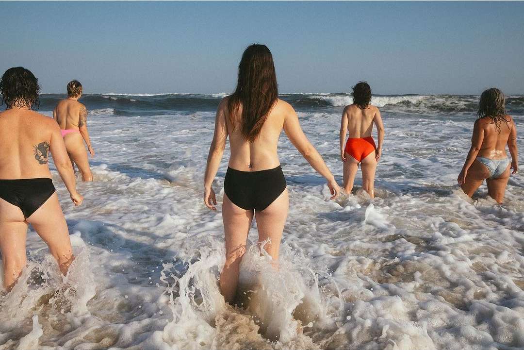 Five women in swimwear stand in ocean surf; beach, waves, and clear sky visible. A group enjoys a refreshing swim in the sea.