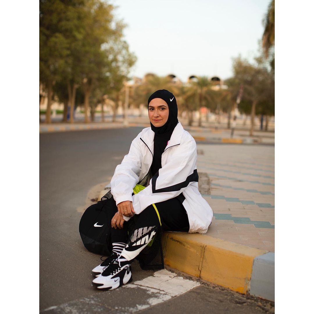 Woman in sportswear sits on curb with Nike shoes and sports bag in outdoor urban setting near trees, wearing hijab and athletic jacket.