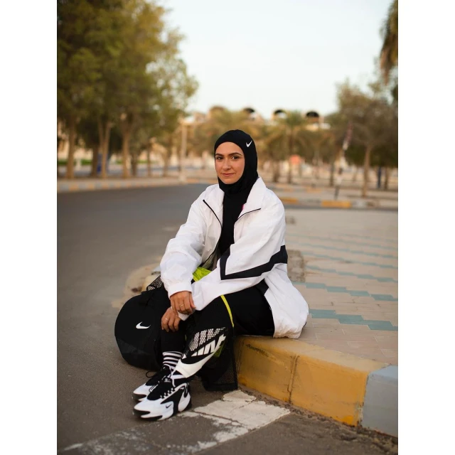 Woman in sportswear sits on curb with Nike shoes and sports bag in outdoor urban setting near trees, wearing hijab and athletic jacket.