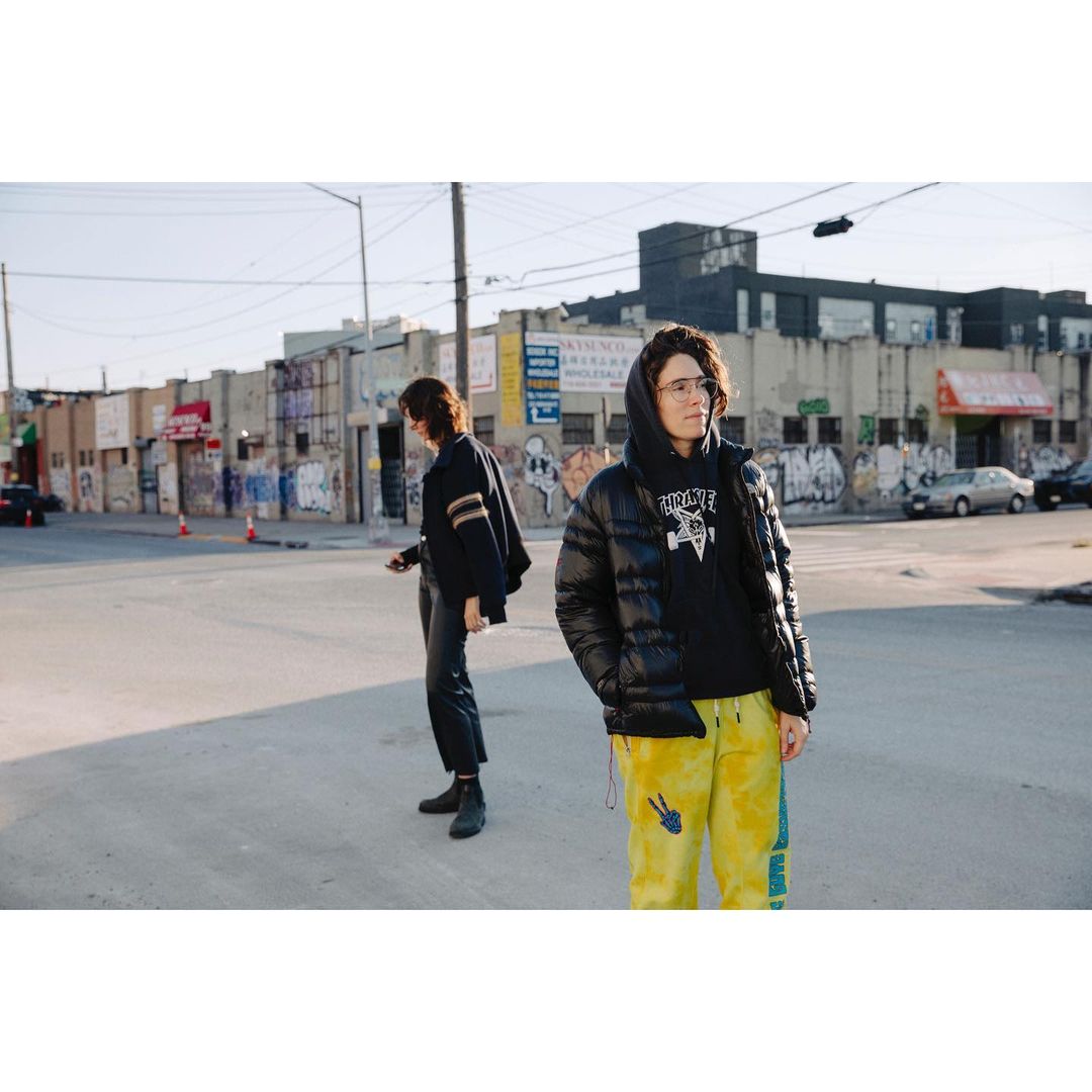 Two people standing on a city street with graffiti-covered buildings and parked cars in the background.
