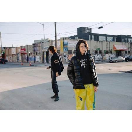 Two people standing on a city street with graffiti-covered buildings and parked cars in the background.