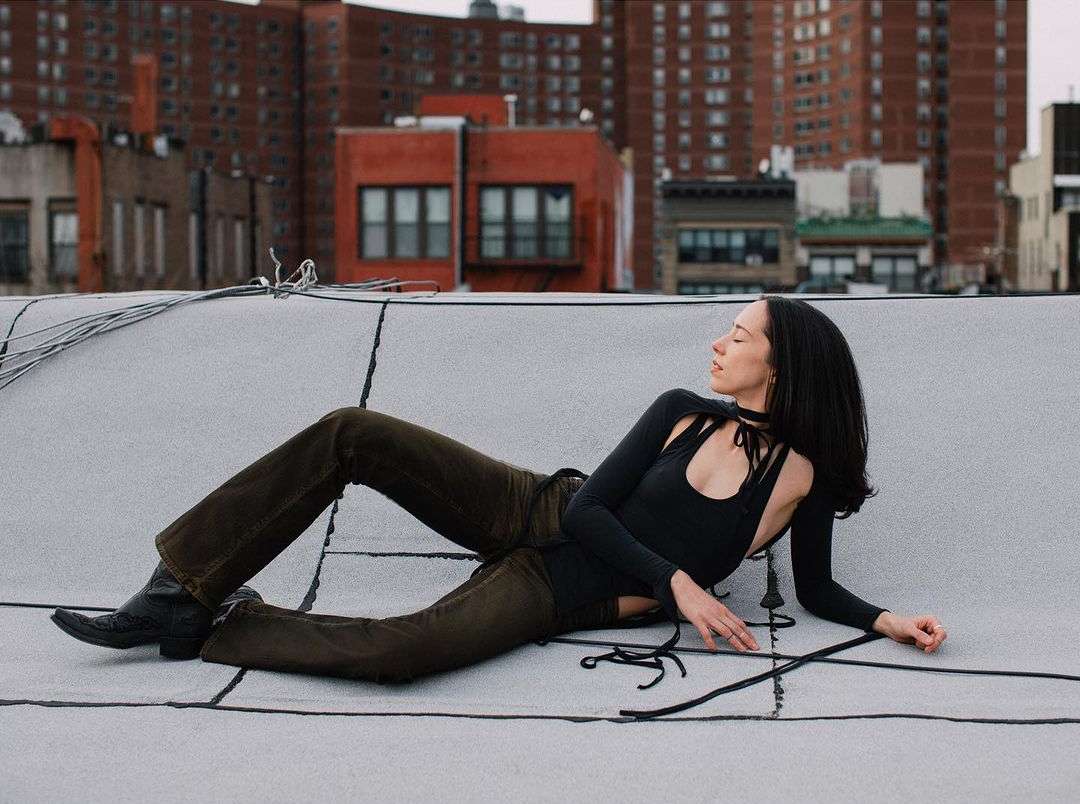 Woman in a black bodysuit and brown pants lying on a rooftop with city buildings in the background, captured in an outdoor fashion shot.