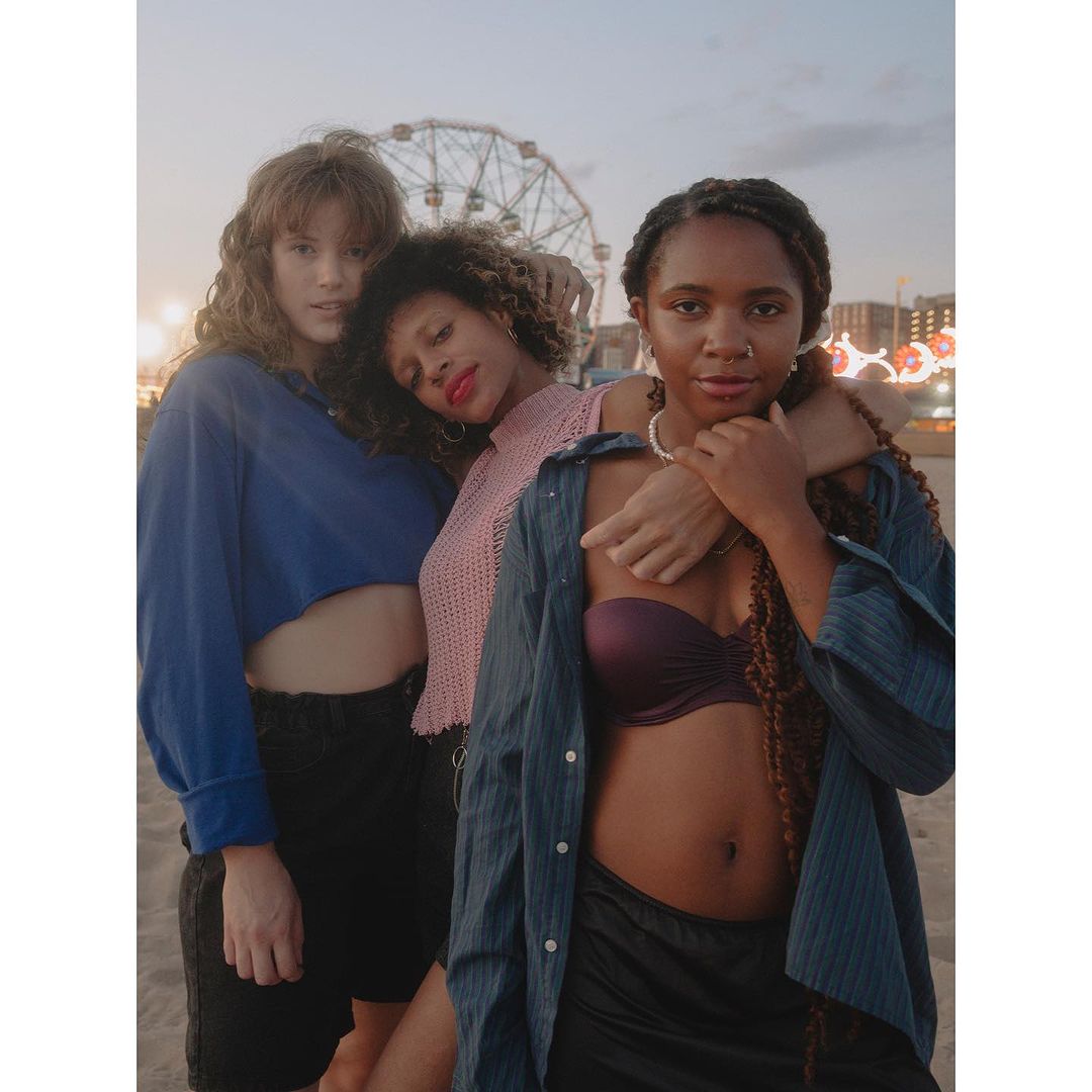 Three young women pose on a beach at dusk, with a Ferris wheel visible in the background. They wear casual tops and shorts, with relaxed expressions.